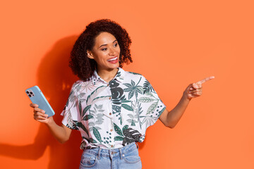 Poster - Photo of brown haired curly girl in summer shirt holding mobile phone pointing finger new eshop isolated on orange color background