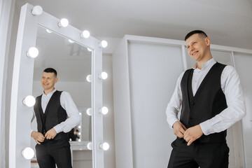 Wall Mural - A man in a black vest stands in front of a mirror, adjusting his tie. The room is well lit, with a large mirror reflecting the man's reflection. The man is in a professional setting