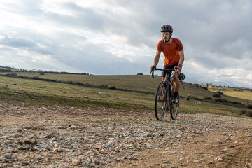 A cyclist rides a gravel bike along a rough gravel path, standing on the pedals as they approach the distant mountains in the background. A dynamic scene of cycling in nature.