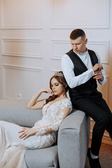 Wall Mural - A bride and groom are sitting on a couch, with the bride wearing a tiara. The man is adjusting the bride's dress