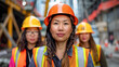 © Kowit - three women of diverse backgrounds wearing hard hats and safety vests at an urban construction site. Position the central figure slightly in front, highlighting her role as a leader