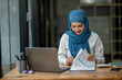 © apichat - A woman wearing a blue scarf is sitting at a desk with a laptop
