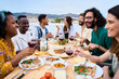 © CarlosBarquero - Gathering of happy people around meal table with wine glasses and food celebration terrace barbecue party. Young multiracial friends enjoying laughing together lunch time on summer weekend on rooftop