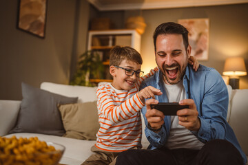 Father and son sit on sofa and use mobile phone at home have fun