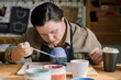 © AnnaStills - Modern young woman with Down syndrome sitting at table coloring handmade ceramic bowl with blue paint