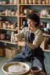 © AnnaStills - Vertical medium long shot of happy Caucasian woman wearing apron sitting at potters wheel shaping clay bowl with her hands