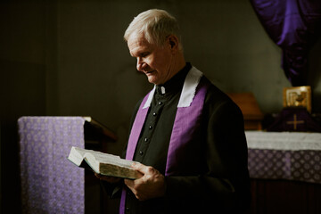 Sticker - Medium shot of elderly Caucasian Catholic pastor wearing soutane holding Bible book reading religious text