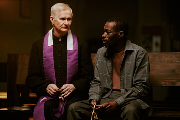African American man holding rosary sitting on pew having conversation with senior Caucasian Catholic priest, medium shot