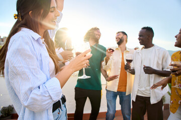 Wall Mural - Young Caucasian beautiful woman holding wine with lens flare, multiracial friends dancing in background on rooftop. Happy generation z people having fun enjoying celebrating summer party together