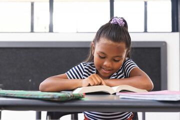 In school, young biracial girl is reading a book at her desk in the classroom