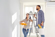 © Studio Romantic - Smiling happy couple painting the wall of their new home holding paint rollers and standing on the ladder. Married man and woman doing repair renovation preparing to move into a new flat.
