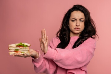 Displeased young dark-haired female showing her food aversion during the studio photo shoot on the pink background. Self-control and nutrition concept