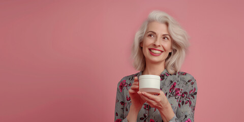 Wall Mural - Mature woman holding white jar with cream on pink background. copy space