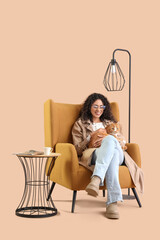 Canvas Print - Young African-American woman with cat sitting in armchair on beige background