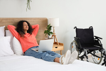 Canvas Print - Young African-American woman with wheelchair and laptop lying on bed at home