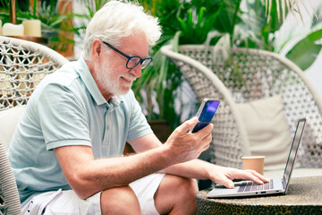 Wall Mural - Portrait of smiling relaxed senior man using mobile phone and laptop sitting on armchair. Old generation people and new technologies