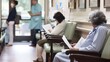 Patients waiting in a hospital lobby, reading documents and filling out paperwork, while medical staff attend to other patients in the background.