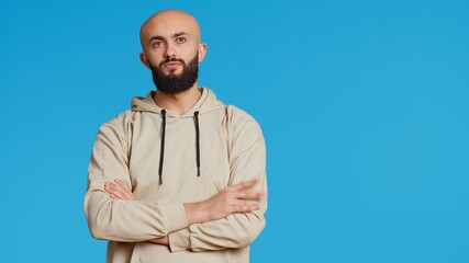 Wall Mural - Young adult in a hoodie waiting for something in studio, standing with arms crossed over blue background. Middle eastern man being impatient on camera, punctual tensed person. Camera 2.