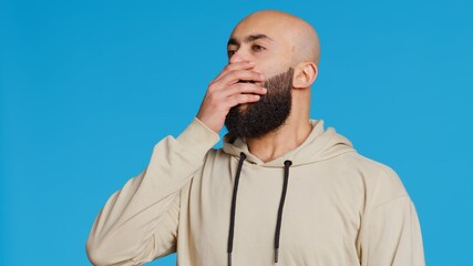 Wall Mural - Middle eastern man feeling exhausted and yawning in studio, covering his mouth in front of the camera. Arab person being sleepy in the morning, awake from the early hours. Camera 2.