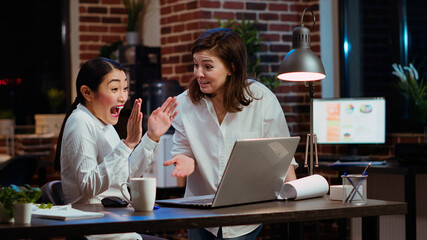 Wall Mural - Jubilant businesswoman and coworker celebrating finalizing deal with customers after working together on laptop. Excited sales consultants doing high five after getting quality leads