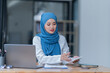 © Tj - A female Arab entrepreneur is diligently working in her office, utilizing her laptop and documents to accomplish her tasks with contentment.