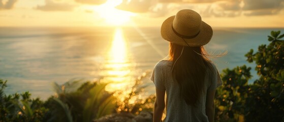 Wall Mural - A young woman, adorned with a hat, overlooks a calm sea in a lush tropical setting. The wide shot captures the serene vibe, bathed in the warm glow of golden hour light.