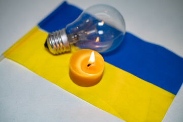 Wall Mural - Burning candle on the background of a light bulb and the Ukrainian flag. Blackout due to the war in Ukraine