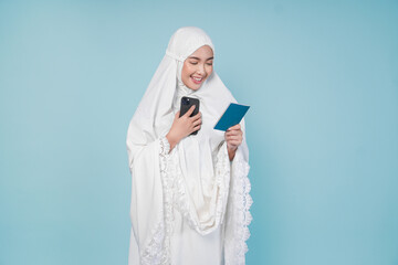 Smiling Asian Muslim woman in prayer gown looking at the passport and smartphone she holds isolated by blue background. Hajj concept.