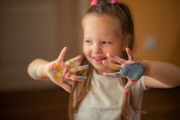 a 5-year-old girl with long blond hair, dressed in a light T-shirt, shows her hands stained with colorful paints, demonstrating her creativity