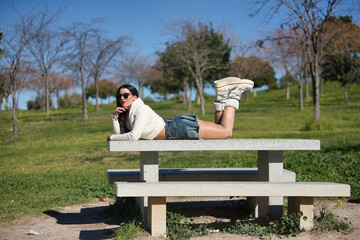 Young, beautiful, brunette woman in a white sweater, denim miniskirt and sunglasses, funny, happy and relaxed, lying on a park bench on a sunny day. Beauty, fashion, trend concept.