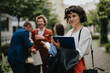 © qunica.com - Young businesswoman holding a clipboard and smiling, while colleagues engage in fun discussion in the background during an outdoor meeting on a sunny day.