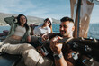 © qunica.com - Group of young adults relaxes while one plays guitar on a boat trip against a scenic mountain backdrop