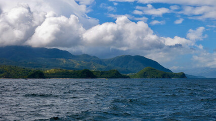 Wall Mural - View from the sea to a tropical island and cumulus clouds. Coastal view of a hilly tropical island and cumulus clouds over a mountain range. Tropical landscape.