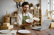 © sofiko14 - Attractive bearded man wearing green apron receiving goods at modern decor store. Professional seller checking for no damages on bowl and plate that holding in hands.