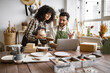 © sofiko14 - Caucasian couple of co-workers work on small pottery production business. Mature man and young curly-haired woman in aprons use laptop to take inventory of products on store website.