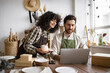 © sofiko14 - Caucasian couple of co-workers work on small pottery production business. Mature man and young curly-haired woman in aprons use laptop to take inventory of products on store website.