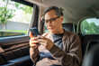 © Chanakon - Relaxing moment of mature man sitting in car back seats using smartphone application on social media look out the window. Senior man happy in car traveling on the road to destination.