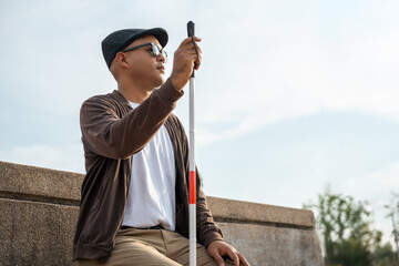 Young asian blind man sitting outside holding walking stick Visually impaired man difficult to traveling on the road wearing sun glasses Cross the road cross the footbridge