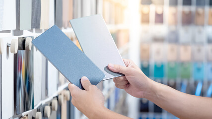 Wall Mural - Designer hands compare two gray laminate sheets nearby material swatch wall display in material library. Choosing color board from collection for interior architecture and furniture finishing design