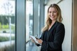 © 为轩 张 - Confident smiling business woman in jacket holding digital tablet in modern office near glass wall. Professional corporate executive leaning against window, looking at camera with a computer