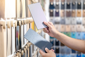 Wall Mural - Designer hands compare two gray laminate sheets nearby material swatch wall display in material library. Choosing color board from collection for interior architecture and furniture finishing design