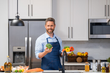 Wall Mural - Middle aged man in cook apron cooking in kitchen. Man on kitchen with vegetables. Portrait of casual man cooking in the kitchen with vegetable ingredients. Guy preparing salad at home in kitchen.