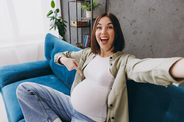 Young pregnant woman wear casual clothes do mobile cell phone selfie shot point on belly sit on blue sofa couch stay home hotel flat rest relax spend free time in living room indoor Maternity concept