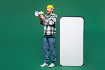 Wall Mural - Full body young man wear shirt blue t-shirt yellow hat big huge blank screen mobile cell phone smartphone with workspace area scream in megaphone isolated on plain green background. Lifestyle concept.