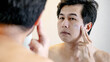 © Summer Paradive - Shirtless Mature Asian man putting cream on his face checking wrinkle in the mirror. Facial complexion treatment and rejuvenation by skincare and cosmetics. Mens beauty cosmetology. Anti aging concept