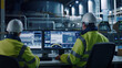 © MYDAYcontent - Two engineers wearing hard hats and safety vests are seated in front of computer monitors, monitoring a manufacturing process in a factory. The monitors display various data and graphs related to the