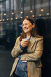 © Maskot - Smiling thoughtful female business professional holding glass and leaning on wall at convention center