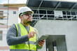 © Ekkasit A Siam - Asian male architect wearing hard hat and reflective vest inspecting construction site with tablet. checking details. Background features unfinished building and construction equipment.