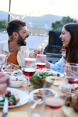 Wall Mural - Vertical. Laughing Caucasian young couple sitting gathered at food table having fun during celebration meal outdoors. Joyful millennial generation happy friends enjoying BBQ on rooftop at sunset