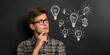 © IBEX.Media - Young man with glasses pondering new ideas, with chalk-drawn light bulbs on a blackboard representing creative thoughts and innovation
